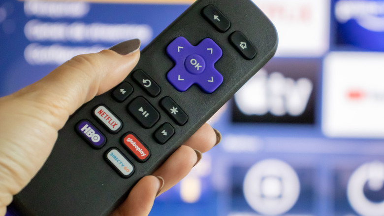 A Roku remote in a woman's hand in front of a smart TV.