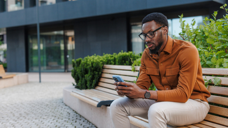A man in glasses sitting on a bench while holding and looking at his phone