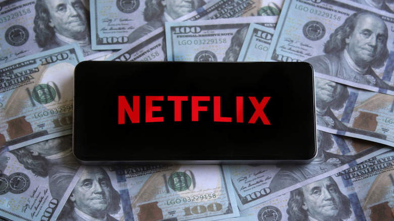 A cellphone displaying the Netflix logo sits atop scatterings of hundred dollar bills, which are covering the background