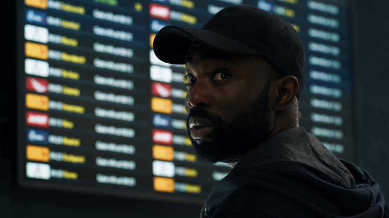 A man in a baseball cap standing in front of an airport flights board, and looking over his shoulder.