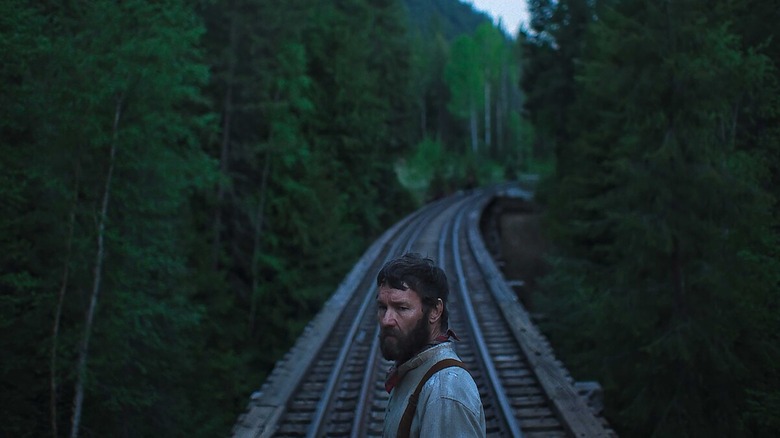 A grizzled man stands on a train track weaving through a lush, green forest.