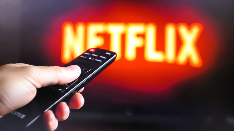 A hand holding a black TV remote in front of a blurred Netflix logo on a TV.
