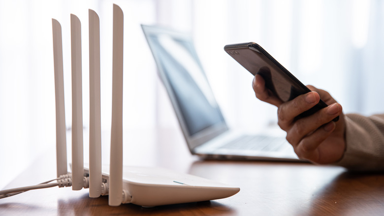 person connecting smartphone to wi-fi router placed on a table with laptop in the background