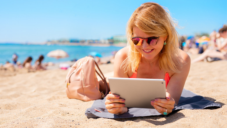 A woman using an iPad while lying on the beach.
