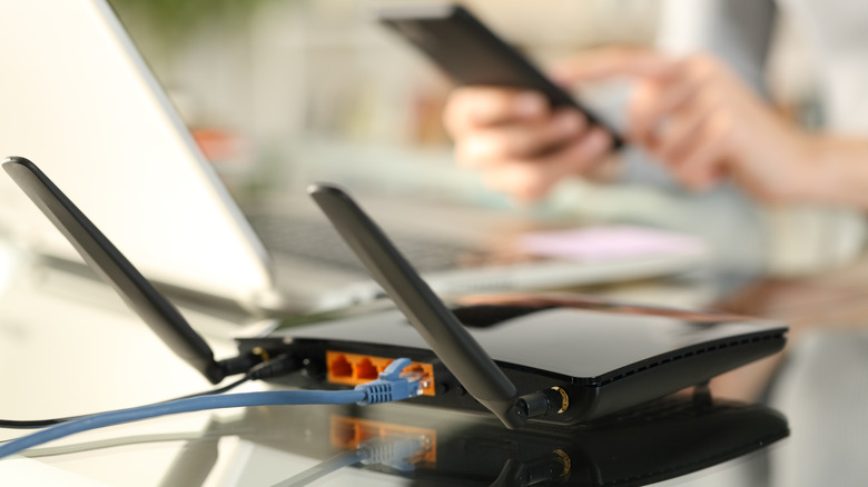 Black router with two antennas with a laptop and person holding a smartphone in background
