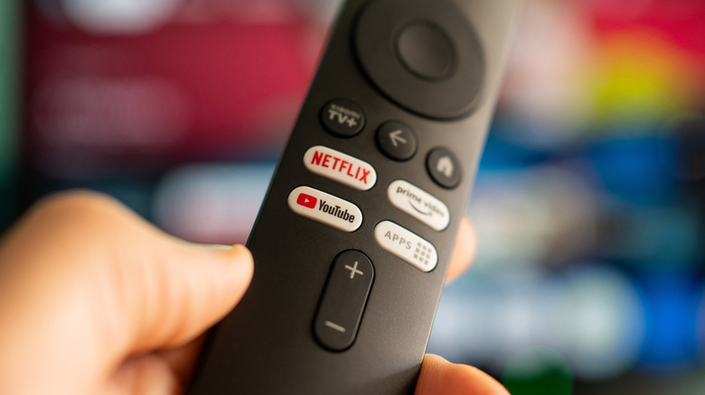 A black remote control held in a user's left hand displays the YouTube logo, among the Netflix Logo and Amazon Prime logo