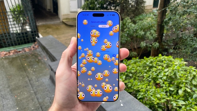 A hand holding an iPhone outdoors. On the screen, the new distorted face emoji