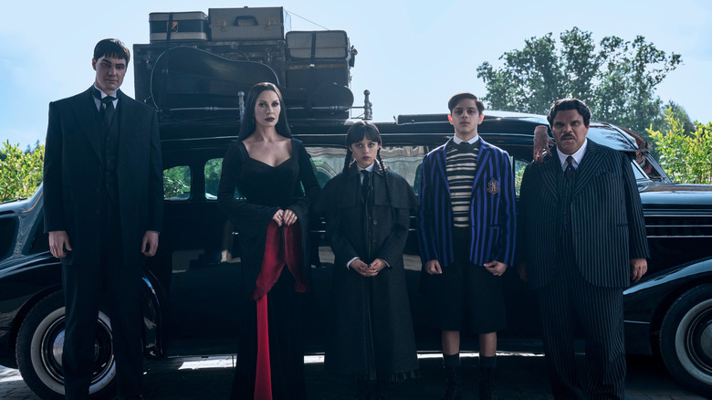 The cast of Wednesday on Netflix stands in front of a car