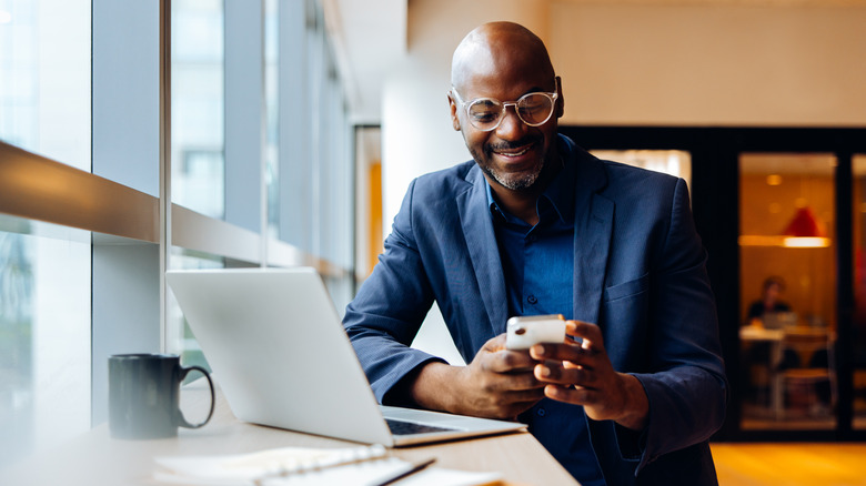 A man in a blue suit and glasses smiles at his phone, while using his computer in a bright work space.