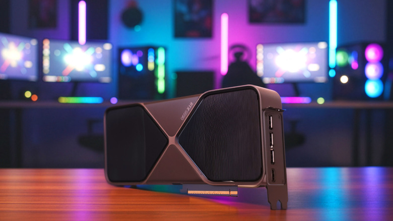 Nvidia RTX 5090 graphics card on a desk, with RGB gaming setup and monitors blurred in the background.