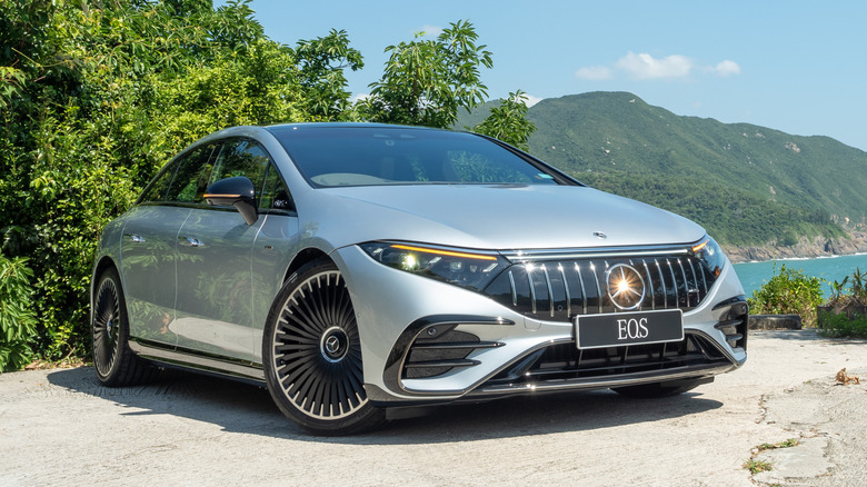 A silver Mercedes-AMG EQS parked in front of lush trees, a large mountain, and a lake