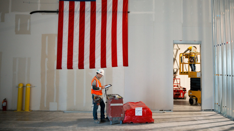 A factory worker looking at a tablet, with the U.S. flag on the wall behind him.