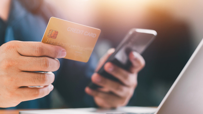 A person in front of a laptop holding a smartphone and credit card.