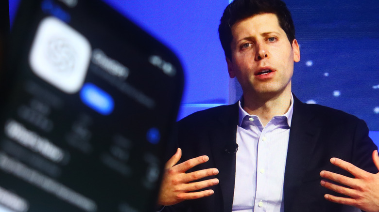 Sam altman motions to himself with his hands, chatGPT icon blurred on a smartphone to the left