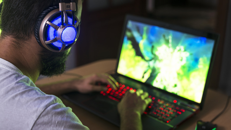 A person wearing a gaming headset using a laptop with its keyboard lit up in red.