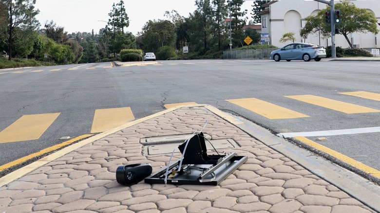 A flock camera lays destroyed in a street median as cars drive by