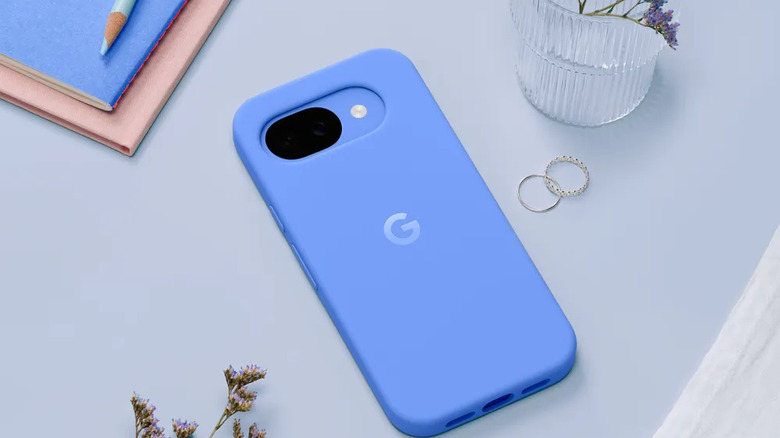 Blue Pixel 10a inside its blue silicone case resting flat on a light blue surface next to rings, a vase with flowers, some thin notebooks and a blue pencil