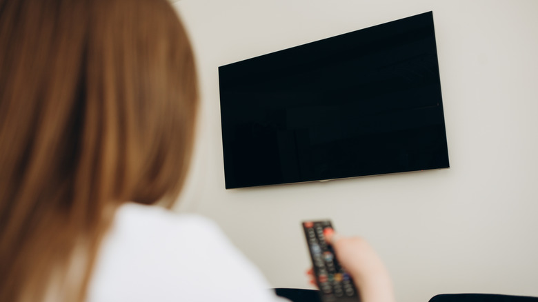 Woman in foreground with remote and a mounted TV on the wall with screen off.