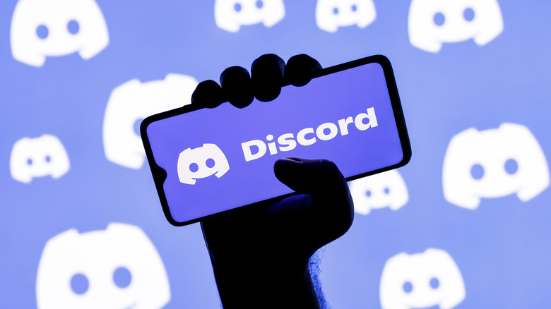 Hand holding up a smartphone with the Discord logo onscreen, and also in the background.