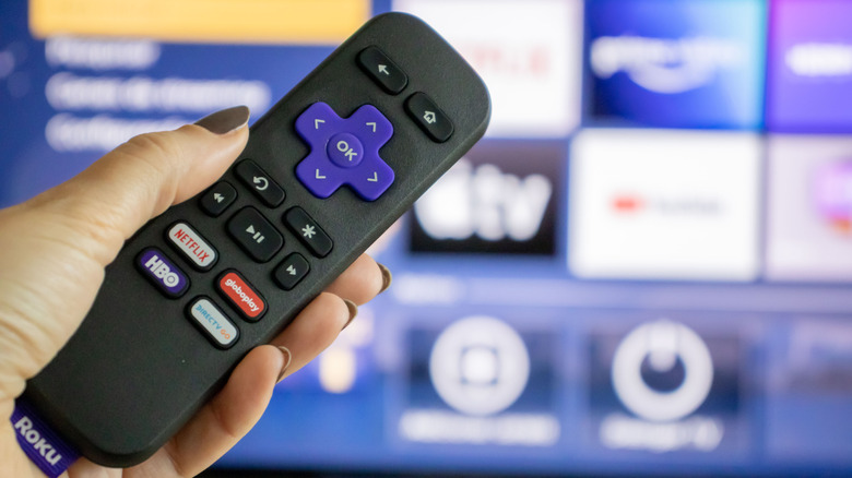 An individual holding a Roku remote in their left hand as it is pointed at a television, the image on the television is blurred but it appears to be the home screen for the Roku operating system