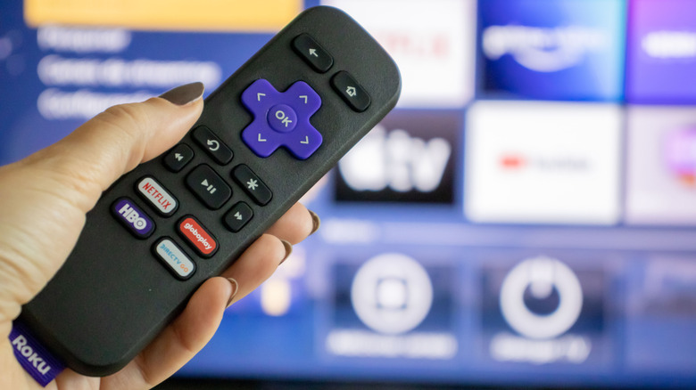 Roku remote in front of blurred Roku user interface on TV.