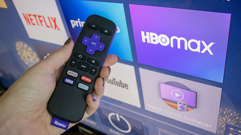 A hand holding a Roku remote with the interface on a display in the background.