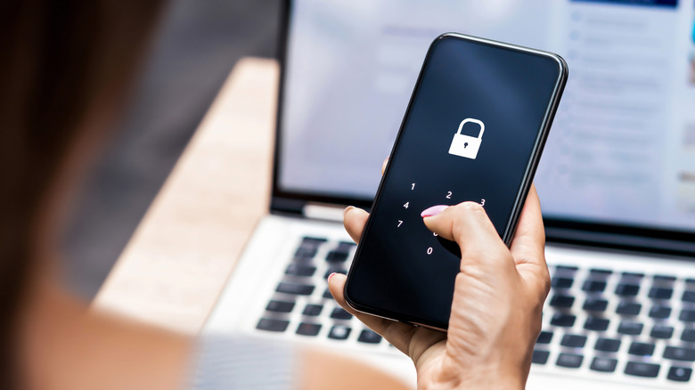 A hand holding a locked phone in front of a laptop.