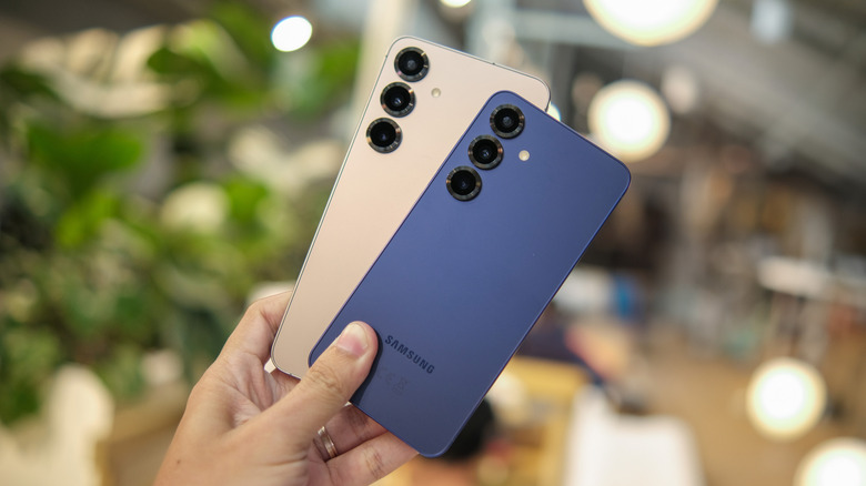 Two Samsung Galaxy S25 smartphones, one blue and one cream, are held in a user's left hand as if they were playing cards, the background is heavily blurred, making it difficult to distinguish what is going on