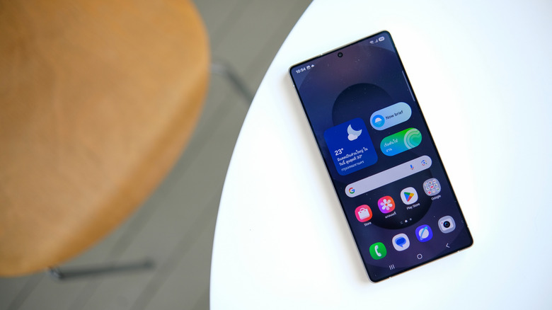A Samsung Galaxy S25 Ultra sits on the edge of a white table, the smartphone is displaying its home screen which contains several apps and widgets