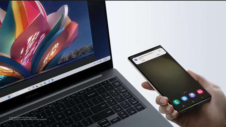 A hand holds a Samsung Galaxy smartphone besides an open Galaxy book laptop. The laptop shows a colorful picture of an abstract flower statue, while the phone receives a notification signifying connectivity.