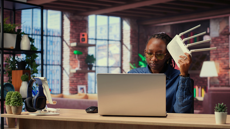 A woman holds a router while typing on a laptop.