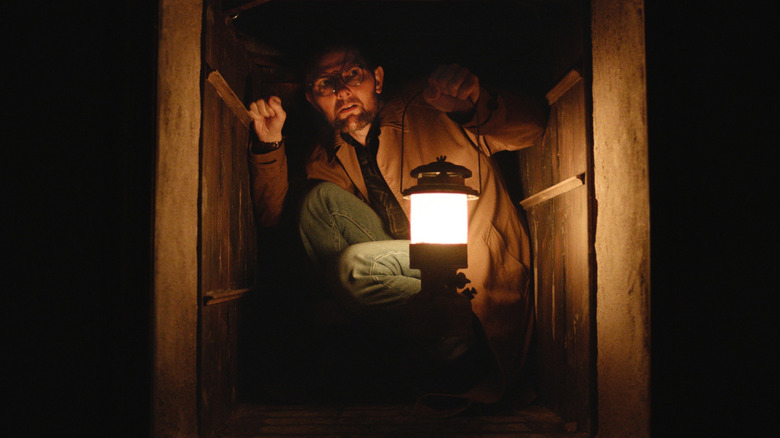 Adam Scott in Hokum, crouched with a lantern in a small space