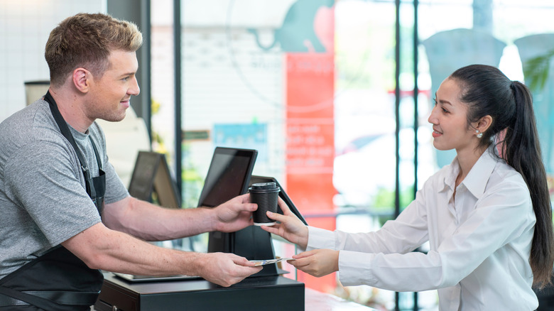A smiling male barista handing a coffee to a female customer as she hands him money