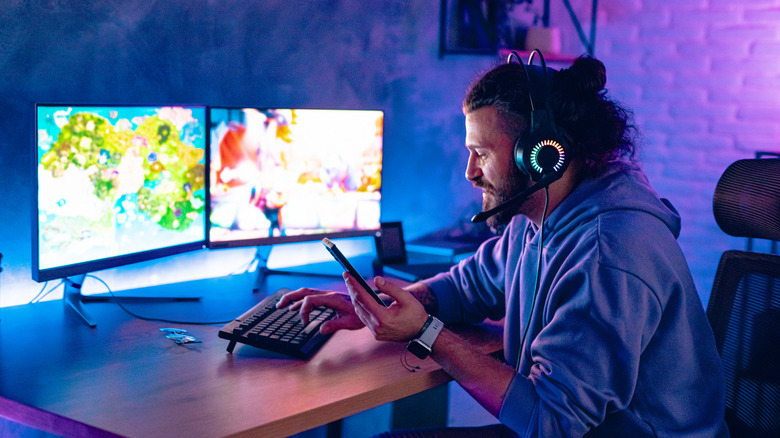 Man wearing a gaming headset while using a phone and keyboard at a dual-monitor desk setup.
