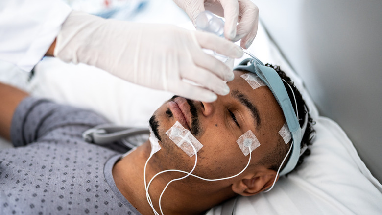 A man lying on a bed while gloved hands attach medial equipment to his head for a sleep study.