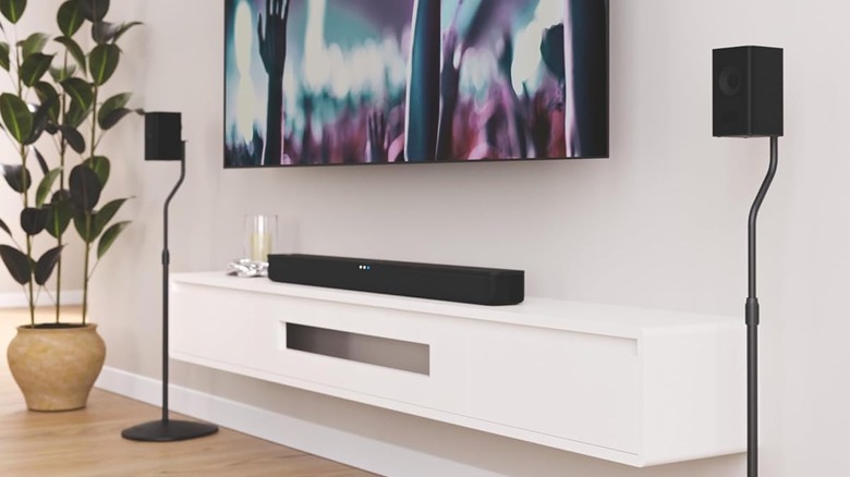 Sanus speaker stands placed on both sides of a wall-mounted TV. The TV is mounted above a wall-mounted AV shelf.