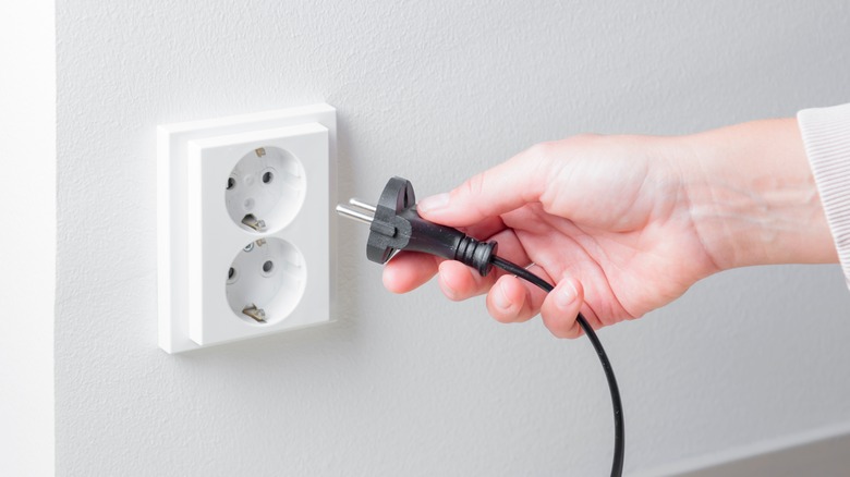 person holding black power cord and plug near white wall outlet