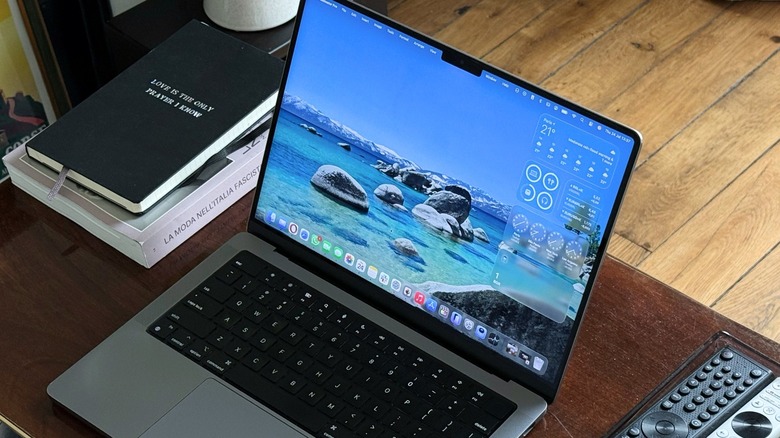 A MacBook Pro on a table with its lid open. There are a few book and remote controls around it.