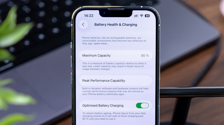 The Battery Health & Charging settings screen on an iPhone within iOS, a MacBook is in the background