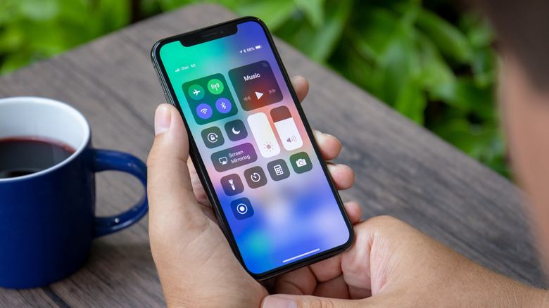 A pair of hands holding an iPhone that is displaying Control Center, there is a table and a coffee cup in the background