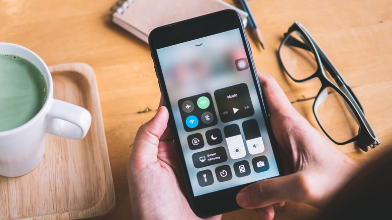 An iPhone displaying Control Center, the device is surrounded by a coffee cup, a pair of glasses, and a notepad