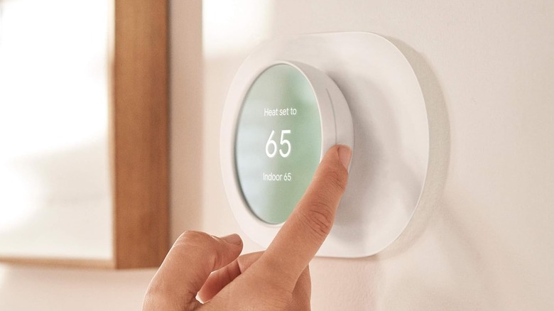 An individual's right index finger is controlling a white Google Nest Smart Thermostat for Home attached to a home wall