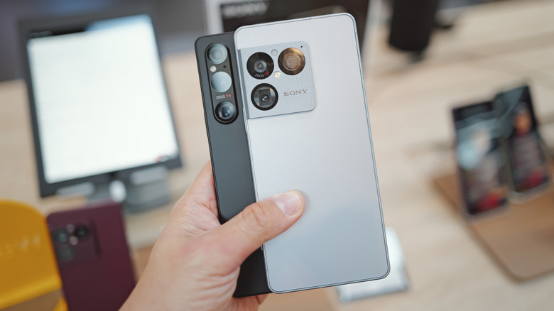 A gray Sony Xperia VIII held in front of a black Xperia V with a shop's mobile display blurred in the background.