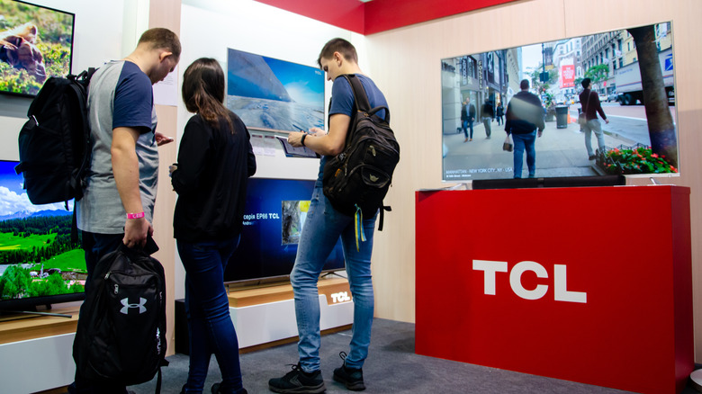 Three individuals stand around a group of TCL televisions attached to the wall, with one television sitting atop a red TCL stand