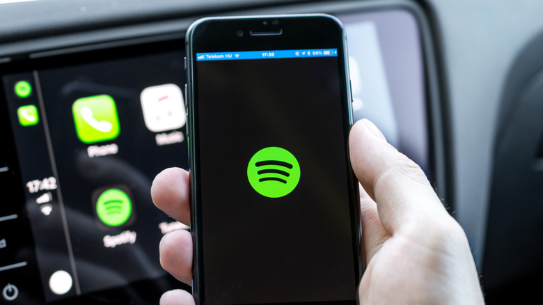 A smartphone displays the Spotify logo as it's held in a user's right hand, behind the device is a vehicle dashboard