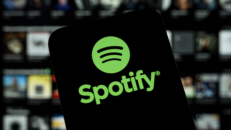 A smartphone displaying the Spotify logo, in the background is a heavily blurred image that appears to be an album selection screen from Spotify