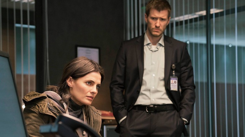 Stana Katic as Emily Byrne sat at a desk and Patrick Heusinger as Nick Durand stood behind her in an office in Absentia
