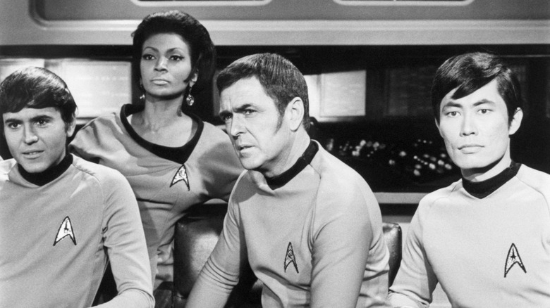 Black and white photo of (from left to right) Checkov, Uhura, Scotty, and Sulu sitting in chairs.