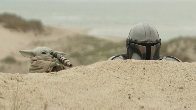 Pedro Pascal as Din Djarin alongside Grogu as they peer over a sand mound in "Star Wars: The Mandalorian and Grogu"