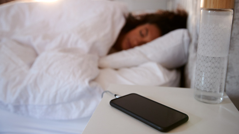 A phone charging on a night stand with a woman sleeping in the background